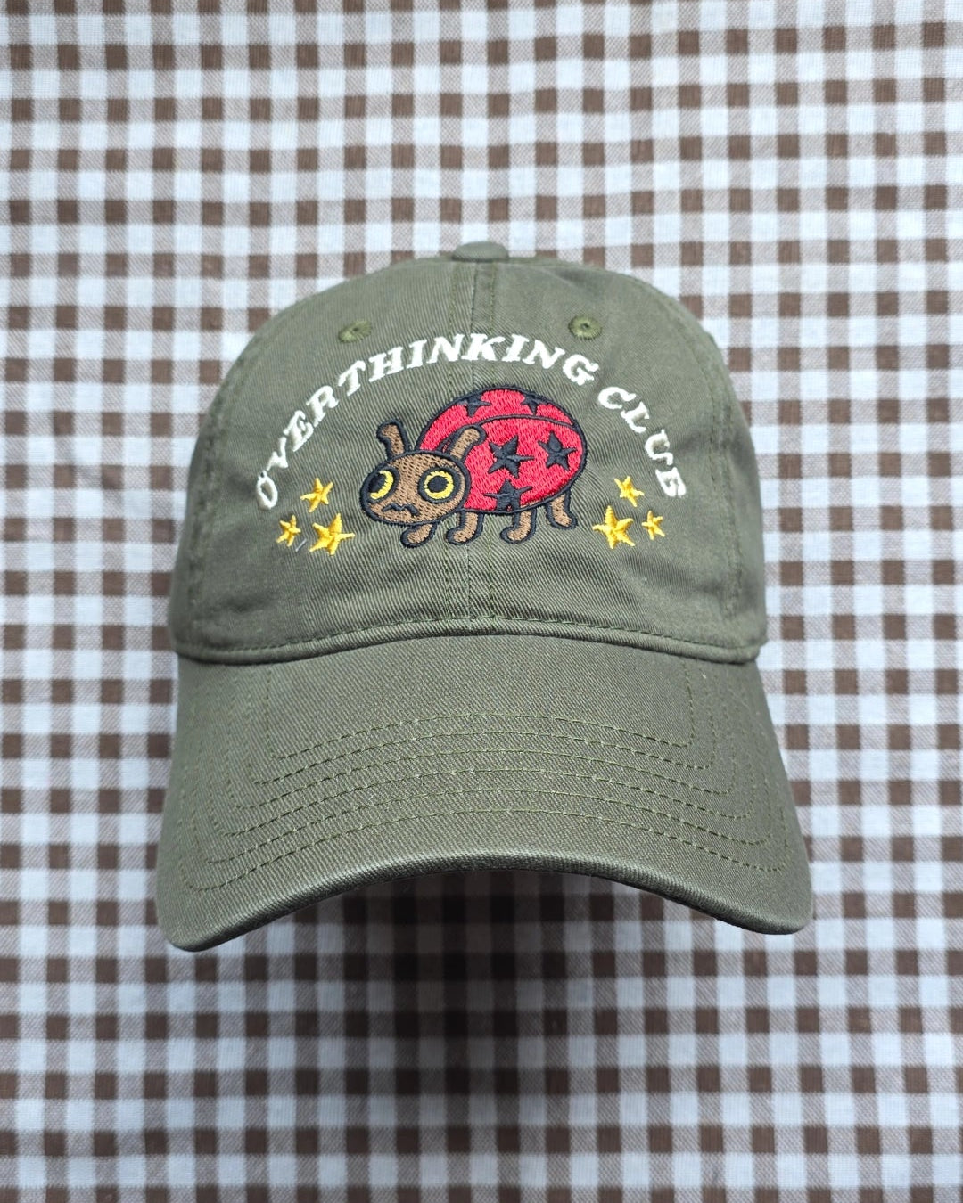 An olive green baseball cap with an embroidered ladybug and the words 'OVERTHINKING CLUB' on the front, displayed on a brown and white checkered fabric background.
